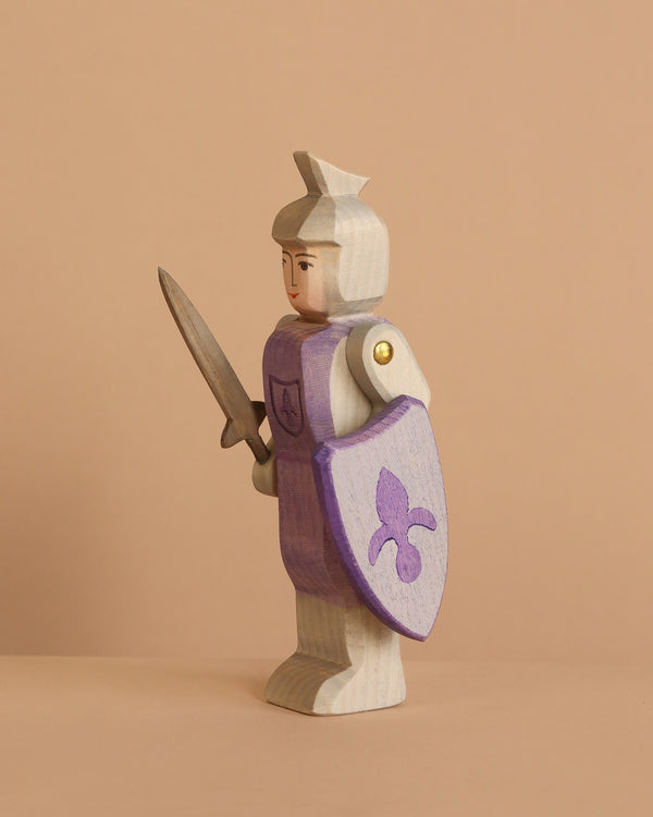 The Ostheimer Blue Knight - Standing is a handcrafted wooden toy with a light blue body, holding a sword and a shield adorned with a blue fleur-de-lis—ideal for imaginative play against any simple backdrop.