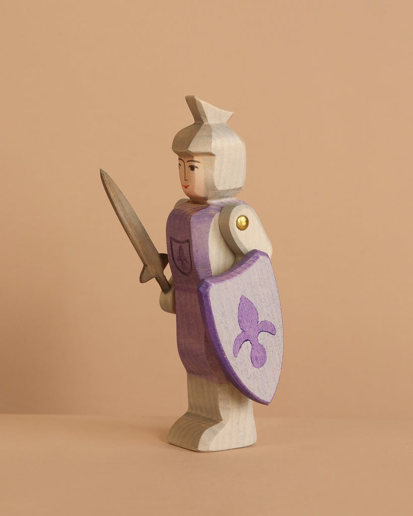 The Ostheimer Blue Knight - Standing is a handcrafted wooden toy with a light blue body, holding a sword and a shield adorned with a blue fleur-de-lis—ideal for imaginative play against any simple backdrop.