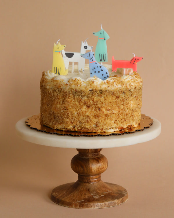 A celebration cake with cream cheese frosting on a wooden cake stand, decorated with colorful Meri Meri Dog Candles in various poses. The background is a soft beige color.