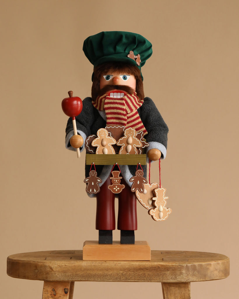 The Christian Ulbricht Collectible Wooden Nutcracker - Gingerbread Vendor Limited Edition 1000 features a festively dressed German nutcracker holding an apple on a stick and gingerbread ornaments, standing on a wooden stool against a beige background.