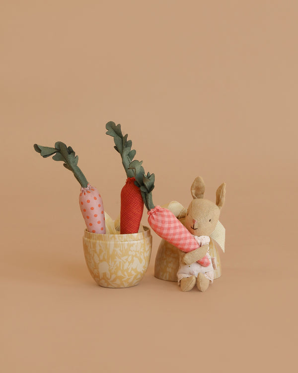 The Maileg Little Rabbit’s Carrot Harvest Set features a petite bunny plush beside a fabric basket filled with three soft carrots. The scene rests against a warm, light tan background for a cozy, minimalist feel.