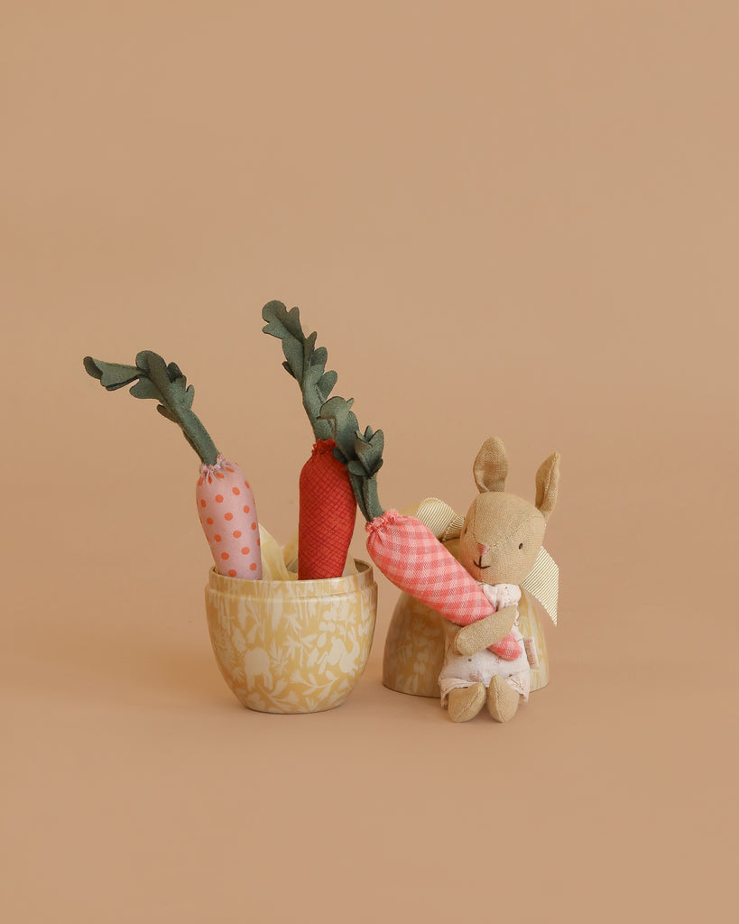 The Maileg Little Rabbit’s Carrot Harvest Set features a petite bunny plush beside a fabric basket filled with three soft carrots. The scene rests against a warm, light tan background for a cozy, minimalist feel.