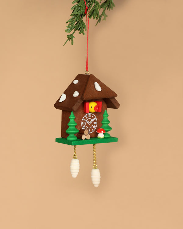 The Christian Ulbricht Cuckoo Clock Ornament, handcrafted in Germany, hangs by a red string. Shaped like a small house with brown roof, green trees, yellow bird, and red mushroom accents, it has cone-shaped weights and small parts that may pose a choking hazard.