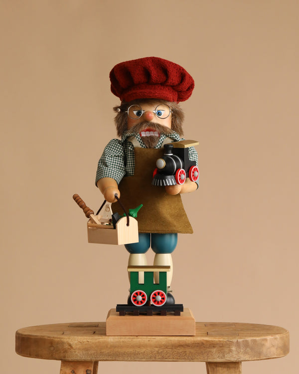 The Christian Ulbricht Collectible Wooden Nutcracker - Railroad Builder Limited Edition 2500 stands on a wooden stool, dressed as a craftsman with a red hat and apron, holding a toy train and toolbox against a beige background.