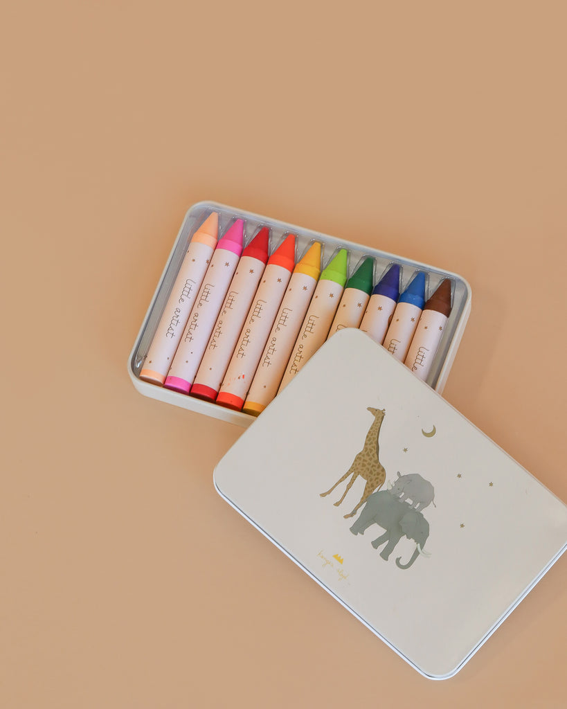 The Konges Sloejd Beeswax Crayons - Safari set features 12 colorful, non-toxic crayons in a tin box with safari animal illustrations. The giraffe and elephant-decorated lid sits beside the open box on a peach background.