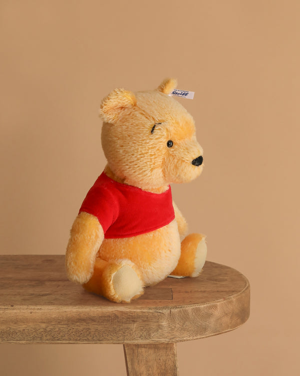 The Steiff Disney’s Winnie the Pooh Open Edition Collectible is a 10-inch plush bear with yellow fur and a red shirt, shown sitting upright on a wooden bench against a plain beige background.