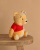 The Steiff Disney’s Winnie the Pooh Open Edition Collectible is a 10-inch plush bear with yellow fur and a red shirt, shown sitting upright on a wooden bench against a plain beige background.