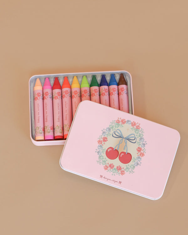 The Konges Sloejd Beeswax Crayons - Cherry Bow come in a pastel pink tin box with floral and cherry designs, holding ten colorful non-toxic crayons neatly arranged inside and visible through the partially open lid.