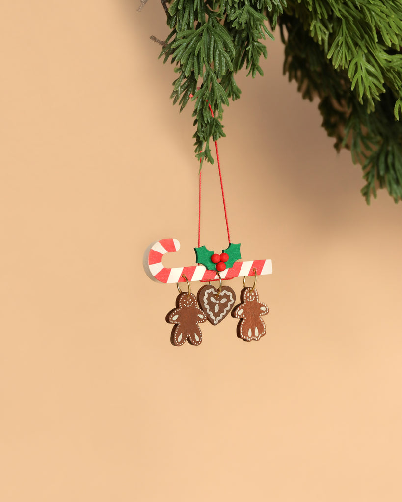 The Christian Ulbricht Candy Cane Ornament, made in Germany, features a festive candy cane adorned with holly leaves and berries at the top, hanging gracefully from a branch. Below the candy cane, two gingerbread figures and a heart shape add charm to this compact ornament set against a warm beige background. It exudes traditional elegance.