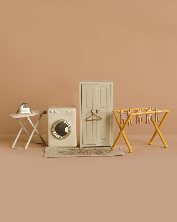 The Maileg Laundry Day Set includes a miniature ironing board with iron, washing machine, door with hanger, and drying rack with clothespins, all displayed against a beige background.