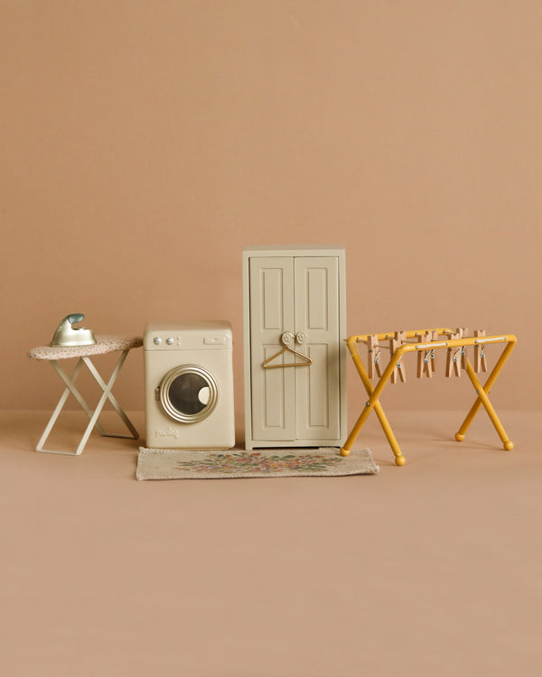 The Maileg Laundry Day Set includes a miniature ironing board with iron, washing machine, drying rack with clothespins, door with hanger, and a small rug—all set against a beige background.