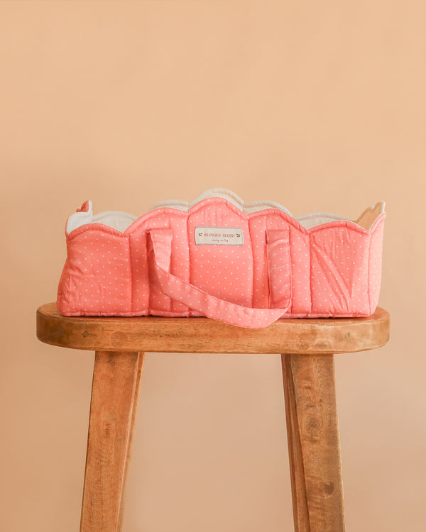 The Konges Sloejd Doll Lift - Rose Dot, a soft pink cotton doll carrier with playful polka dots and a convenient handle, rests atop a wooden stool against a peach-colored backdrop. This padded doll lift is OEKO-TEX certified, providing safe and comfortable transport for your little one’s favorite toys.