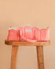 The Konges Sloejd Doll Lift - Rose Dot, a soft pink cotton doll carrier with playful polka dots and a convenient handle, rests atop a wooden stool against a peach-colored backdrop. This padded doll lift is OEKO-TEX certified, providing safe and comfortable transport for your little one’s favorite toys.