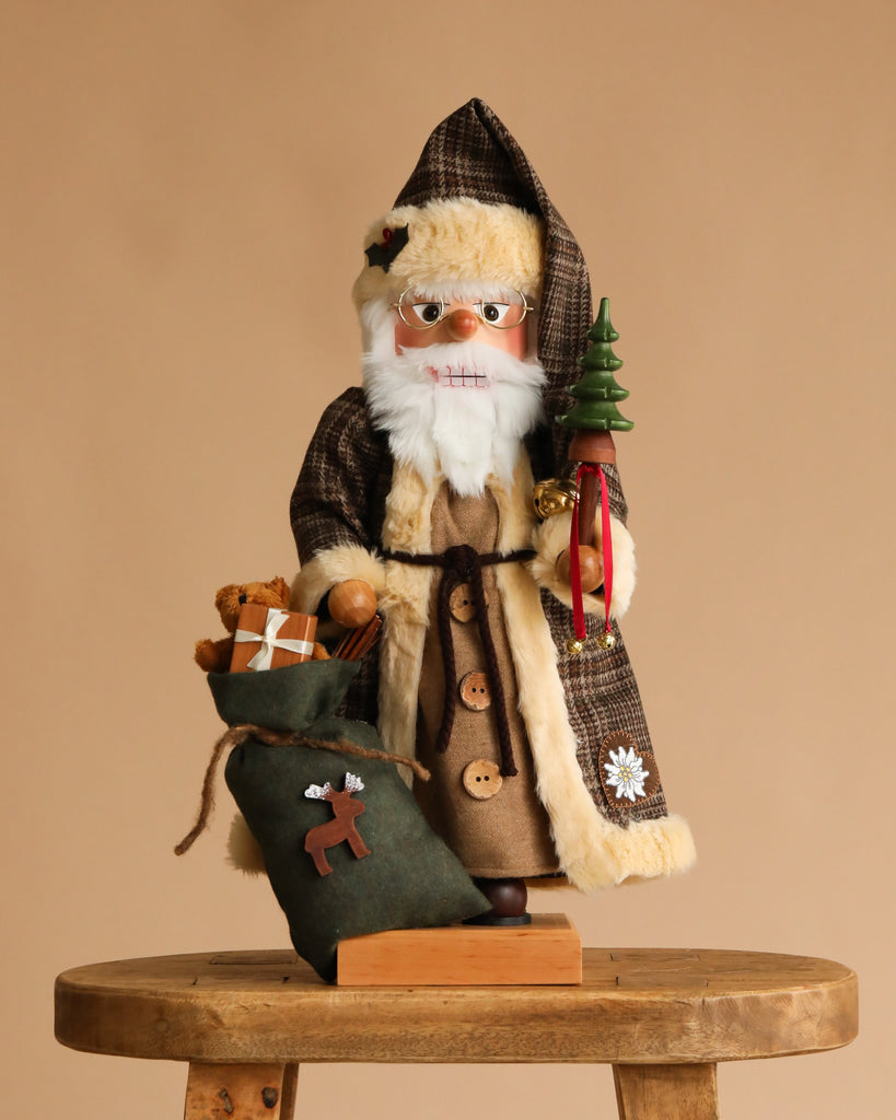 The Christian Ulbricht Collectible Wooden Nutcracker features Santa Claus in a brown checkered coat, standing on a stool with a green Christmas tree, green gift sack, and teddy bear, set against a plain beige background.