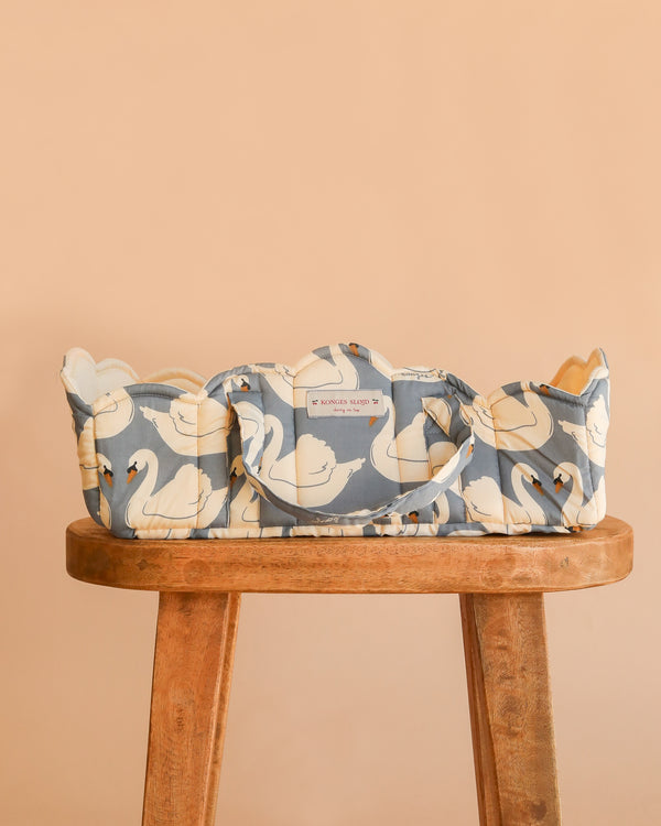 The Konges Sloejd Doll Lift - Swan, crafted from OEKO-TEX certified cotton and adorned with a blue and white swan pattern, sits atop a wooden stool against a peach background. Offering both style and practicality for playtime, this charming basket ensures safety and comfort for your little one’s favorite toys.