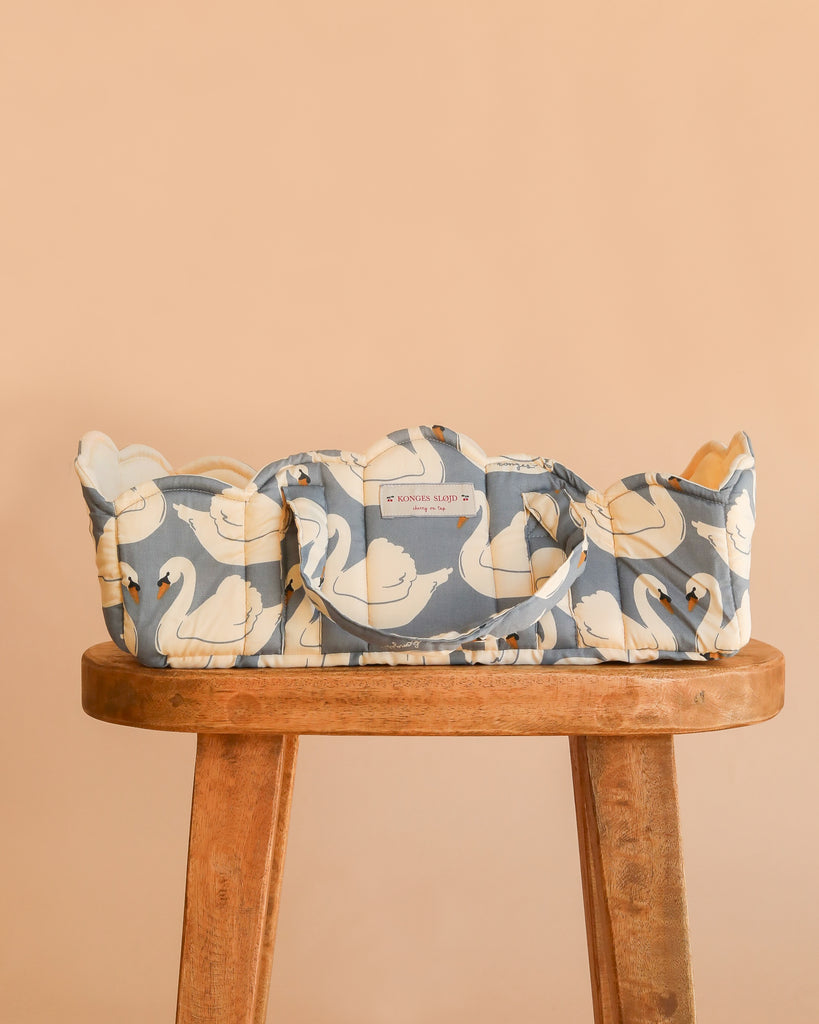 The Konges Sloejd Doll Lift - Swan, crafted from OEKO-TEX certified cotton and adorned with a blue and white swan pattern, sits atop a wooden stool against a peach background. Offering both style and practicality for playtime, this charming basket ensures safety and comfort for your little one’s favorite toys.