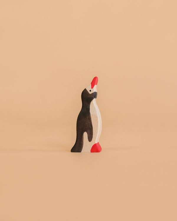 A Holztiger Penguin, Small, Head Raised—handcrafted from wood and featuring black, white, and red accents—stands upright on a plain peach-colored background.