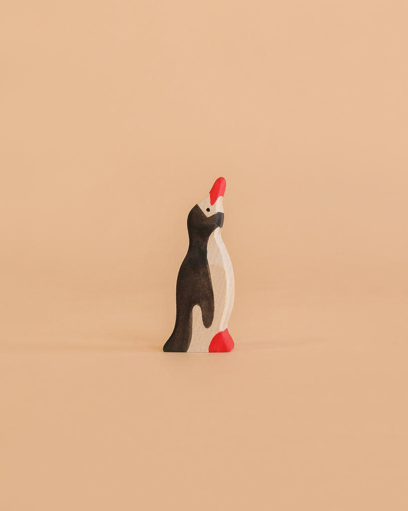 A Holztiger Penguin, Small, Head Raised—handcrafted from wood and featuring black, white, and red accents—stands upright on a plain peach-colored background.