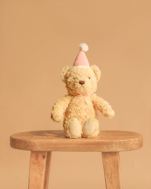 The Snuggle Bunnies - Birthday Arthur - Fairy, a light brown plush bear in a pink party hat with a white pom-pom, sits on a wooden stool against a simple beige background.
