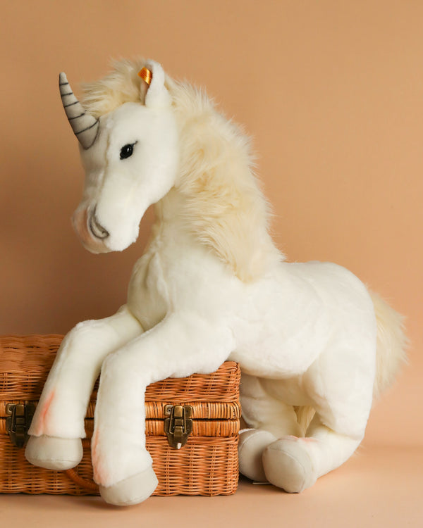 A large white Steiff, Starly Unicorn Plush Animal Toy, 28 Inches with a fluffy mane and a silver horn is leaning on a wicker basket. This premium-grade woven plush features a soft expression and is set against a warm beige background, embodying the quality of handmade Steiff craftsmanship.