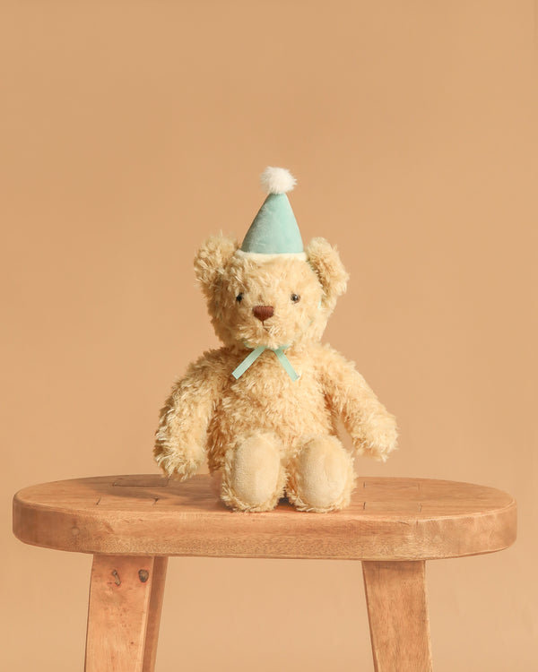 Snuggle Bunnies - Birthday Arthur - Sky is a plush beige teddy bear with a light blue party hat and ribbon, seated on a wooden stool against a tan background—a charming keepsake gift for birthdays.