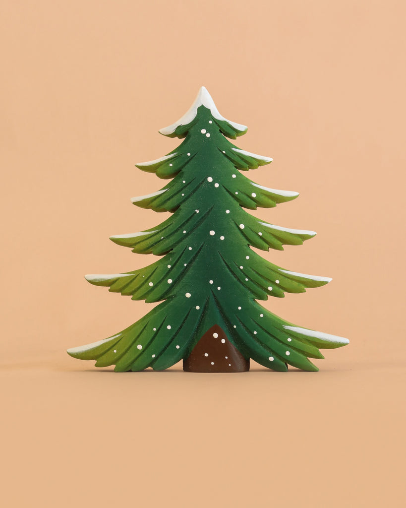 The Bumbu Handmade Wooden Snowy Fir Tree, Large, is a Montessori-inspired, eco-friendly wooden toy featuring stylized green branches accented with white snow, showcased against a plain peach background.