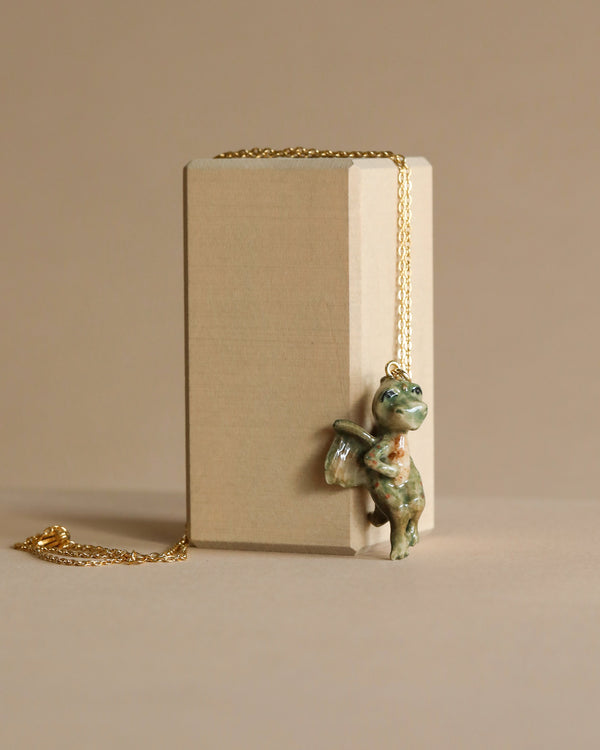 A handcrafted porcelain Baby Dragon Necklace with delicate painting details hangs from a gold chain draped over a plain beige book on a matching background.