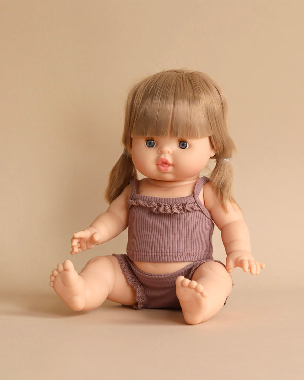 A Minikane Baby Doll (13") - Yzé with blonde hair styled in two pigtails sits on a neutral background. The doll is wearing a matching purple tank top and bottoms. Its blue eyes and slightly open mouth give it a lifelike appearance. The doll's anatomically correct hands rest on the ground, and its feet are bare. The delicate fragrance of natural vanilla scent adds to its charm.