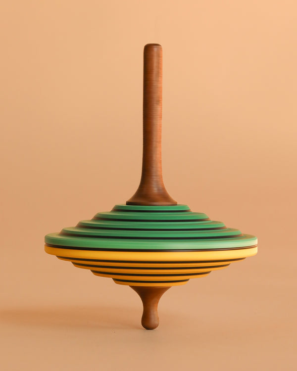 The Mader Tukan Spinning Top - Green & Yellow is a wooden toy with layered disc-like sections in brown, green, yellow, and black, standing upright against a matching beige background.