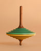 The Mader Tukan Spinning Top - Green & Yellow is a wooden toy with layered disc-like sections in brown, green, yellow, and black, standing upright against a matching beige background.