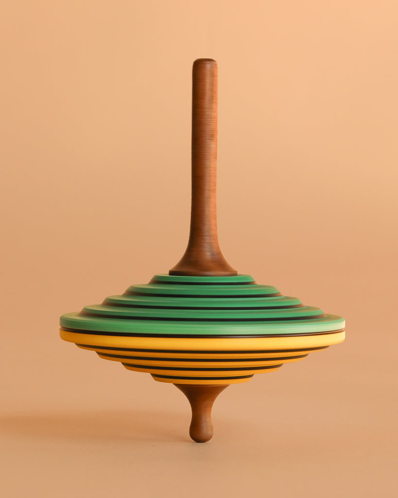 The Mader Tukan Spinning Top - Green & Yellow is a wooden toy with layered disc-like sections in brown, green, yellow, and black, standing upright against a matching beige background.