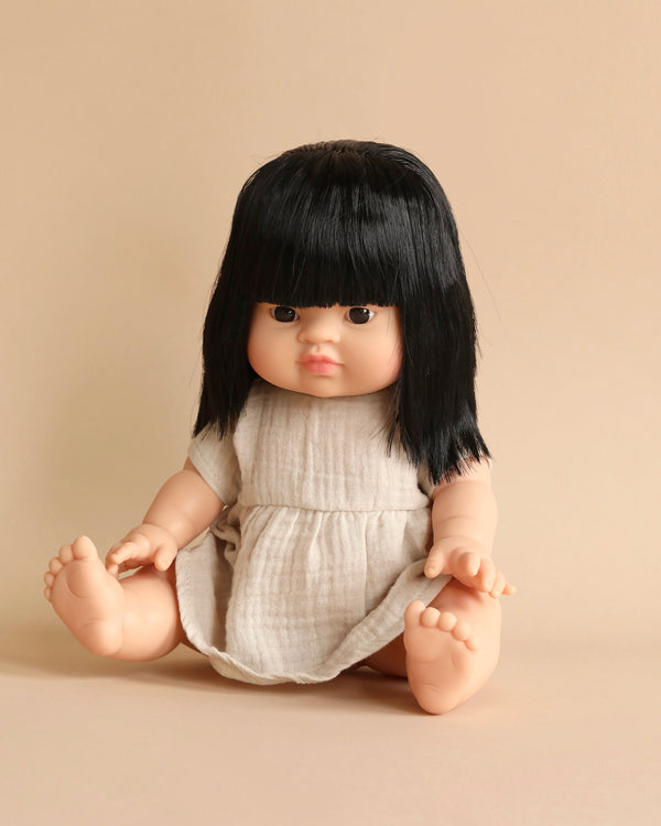 A Minikane Baby Doll (13") - Jade with long black hair and bangs is seated against a plain beige background. The doll, known for its anatomically correct features, wears a simple light-colored dress and has a neutral expression, with its arms and legs slightly apart.