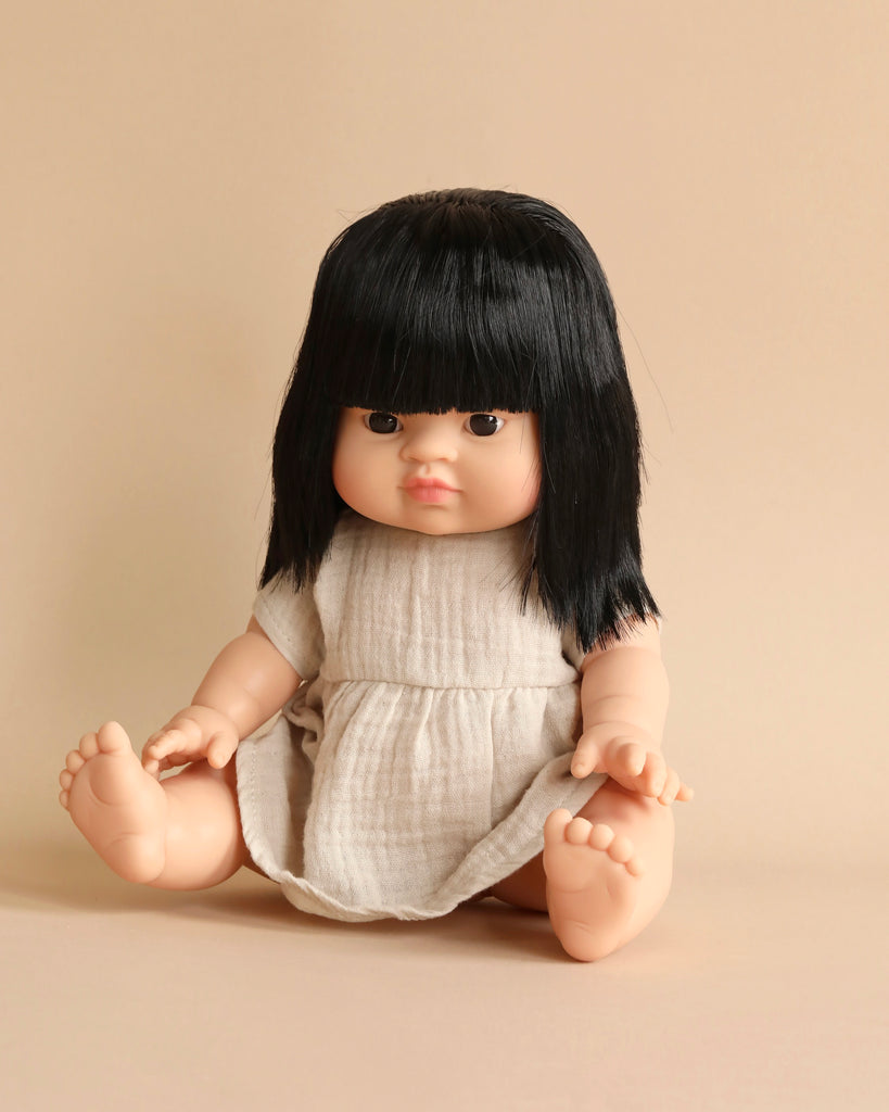 A Minikane Baby Doll (13") - Jade with long black hair and bangs is seated against a plain beige background. The doll, known for its anatomically correct features, wears a simple light-colored dress and has a neutral expression, with its arms and legs slightly apart.