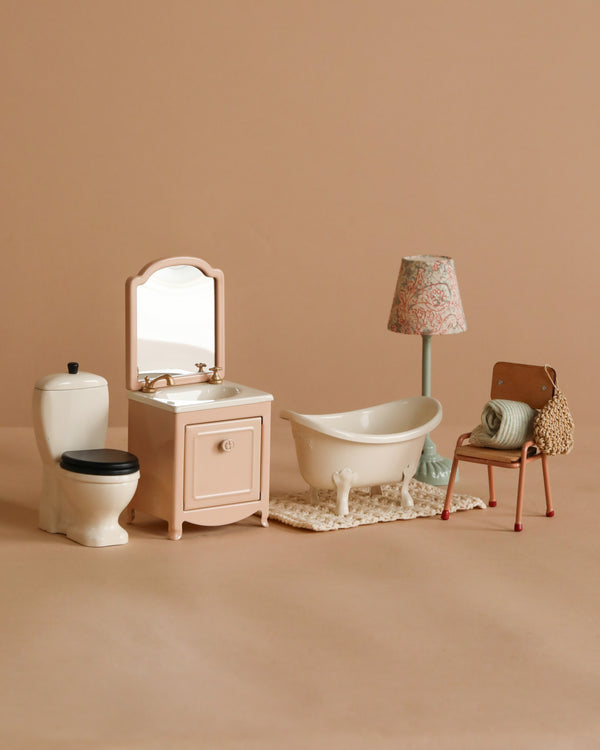 The Maileg Bathroom Set includes a bathtub, toilet, sink with mirror, clawfoot tub on a rug, floral floor lamp, and chair with towel and crocheted bag—perfect accessories for your Maileg Castle.