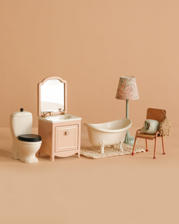 The Maileg Bathroom Set includes a bathtub on a rug, toilet, sink with mirror, floral floor lamp, chair with folded towels, and a small bag, all artfully displayed against a beige background.
