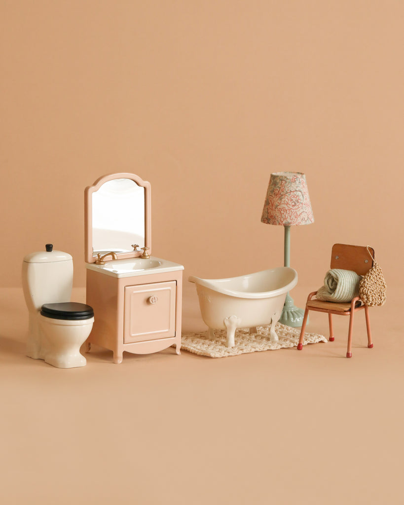 The Maileg Bathroom Set includes a bathtub on a rug, toilet, sink with mirror, floral floor lamp, chair with folded towels, and a small bag, all artfully displayed against a beige background.