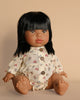 A Minikane Baby Doll (13") - Latika with long black hair is positioned against a beige background. The doll wears a light-colored romper featuring a floral pattern and buttons down the front. It showcases detailed facial features, legs extended forward, and is accompanied by a natural vanilla scent.