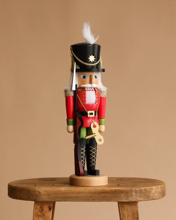 The Christian Ulbricht Collectible Wooden Nutcracker - Toy Soldier features a black hat, red and green uniform, and white hair, standing on a wooden stool against a plain beige background.