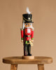 The Christian Ulbricht Collectible Wooden Nutcracker - Toy Soldier features a black hat, red and green uniform, and white hair, standing on a wooden stool against a plain beige background.