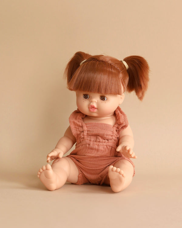 A seated Minikane Baby Doll With Sleeping Eyes (13") - Raphaëlle with light skin, brown eyes, and short, brown hair styled in pigtails. The anatomically correct doll is wearing a pale brown romper and is posed against a matching light brown background. It also features a natural vanilla scent for added charm.