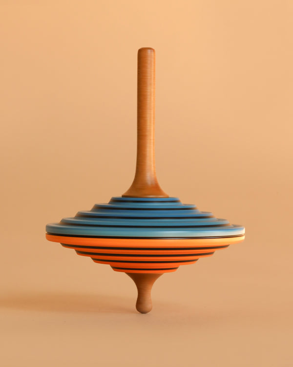 The Mader Tukan Spinning Top - Blue & Orange is a handcrafted Austrian toy with layered blue, orange, black, and natural wood rings. It stands upright and adds a colorful touch against a light beige background.