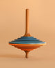 The Mader Tukan Spinning Top - Blue & Orange is a handcrafted Austrian toy with layered blue, orange, black, and natural wood rings. It stands upright and adds a colorful touch against a light beige background.