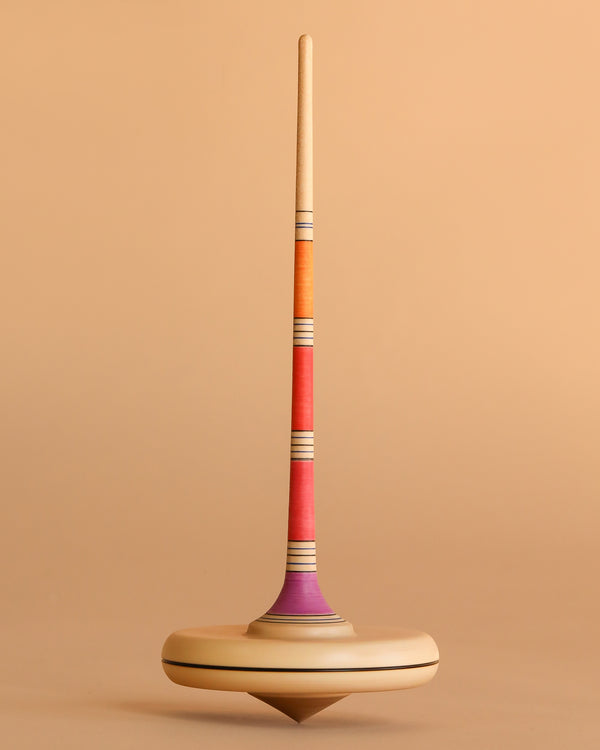 The Mader Arabella Spinning Top - Pink is a wooden toy with a tall, slender handle and vibrant orange, red, and purple stripes, shown upright against a light brown background—reflecting Arabella’s commitment to safe, colorful design.