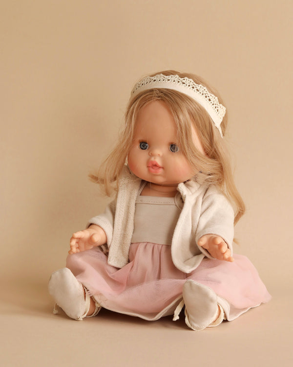 A Minikane Baby Doll (13") - Princess Eleanor with long blonde hair sits against a beige background, wearing a cream-colored headband, a light pink tulle skirt, a cream sweater, and matching soft shoes. The anatomically correct doll features a neutral expression with closed lips and slightly open eyes.