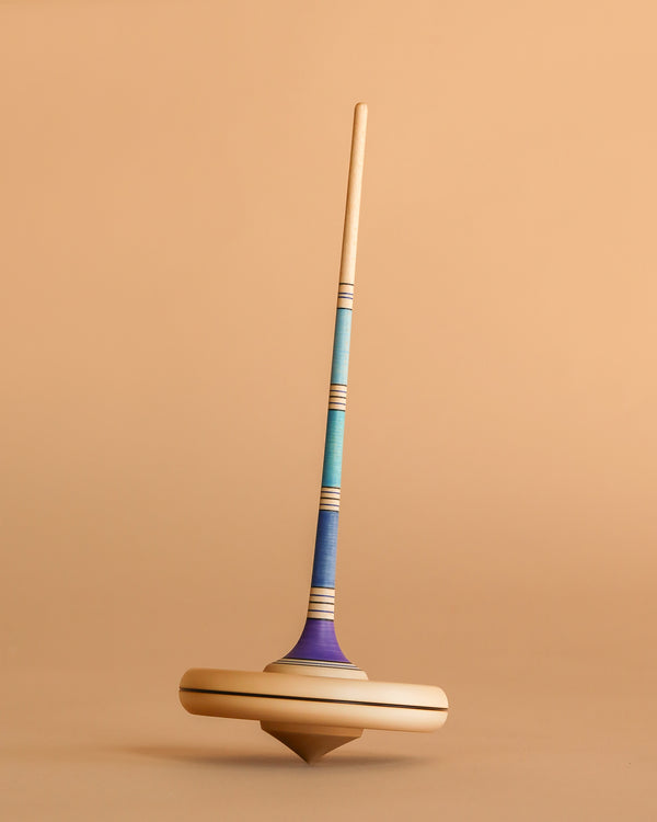The Mader Arabella Spinning Top - Blue is a wooden toy with a long handle and blue, teal, and purple stripes, standing upright on a tan surface against a matching background.