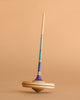 The Mader Arabella Spinning Top - Blue is a wooden toy with a long handle and blue, teal, and purple stripes, standing upright on a tan surface against a matching background.