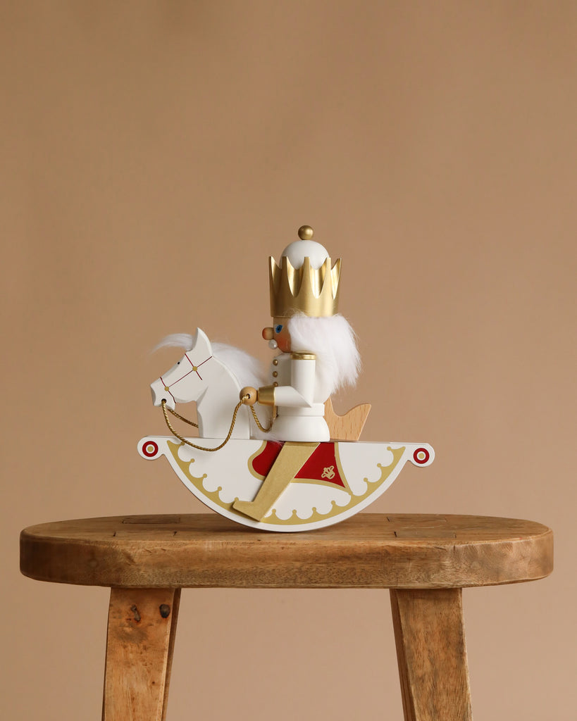 The Christian Ulbricht Collectible Wooden Nutcracker - Rider King, wearing a crown and gold-red accents, sits on a white rocking horse atop a wooden stool against a plain beige background.