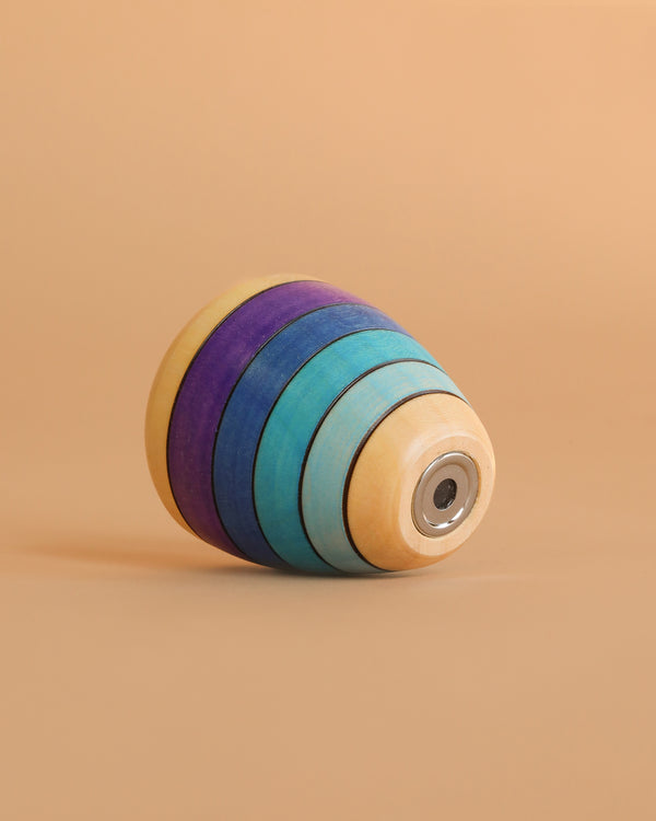 The Mader Wooden Kaleidoscope - Purple is a handcrafted ash spinning top with yellow, blue, teal, and purple concentric stripes, shown lying on its side against a beige background.