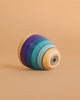 The Mader Wooden Kaleidoscope - Purple is a handcrafted ash spinning top with yellow, blue, teal, and purple concentric stripes, shown lying on its side against a beige background.