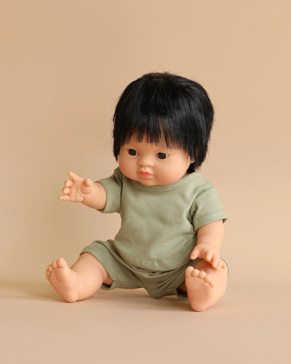 A Minikane Baby Doll (13") - Jude with black hair wearing a light green shirt and shorts is sitting against a beige background. The anatomically correct doll has its right arm extended forward and its legs stretched out on the floor.